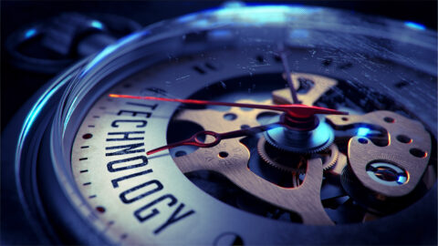 Close-up of a precision mechanical timepiece with visible gears, polished metallic components, glass casing, and the word 