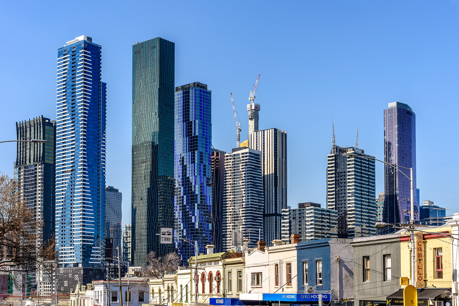 Melbourne city skyline in Victoria, Australia, featuring modern skyscrapers and buildings under construction rising above older North Melbourne buildings under a clear blue sky.