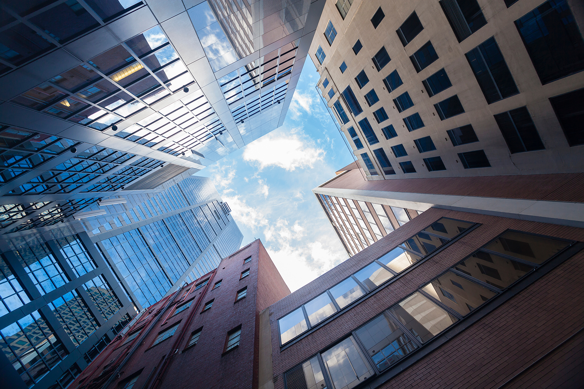 Looking upward between modern skyscrapers in Melbourne CBD, Australia, with glass and brick commercial buildings surrounding a bright blue sky, representing facility management and unsupervised building operations.