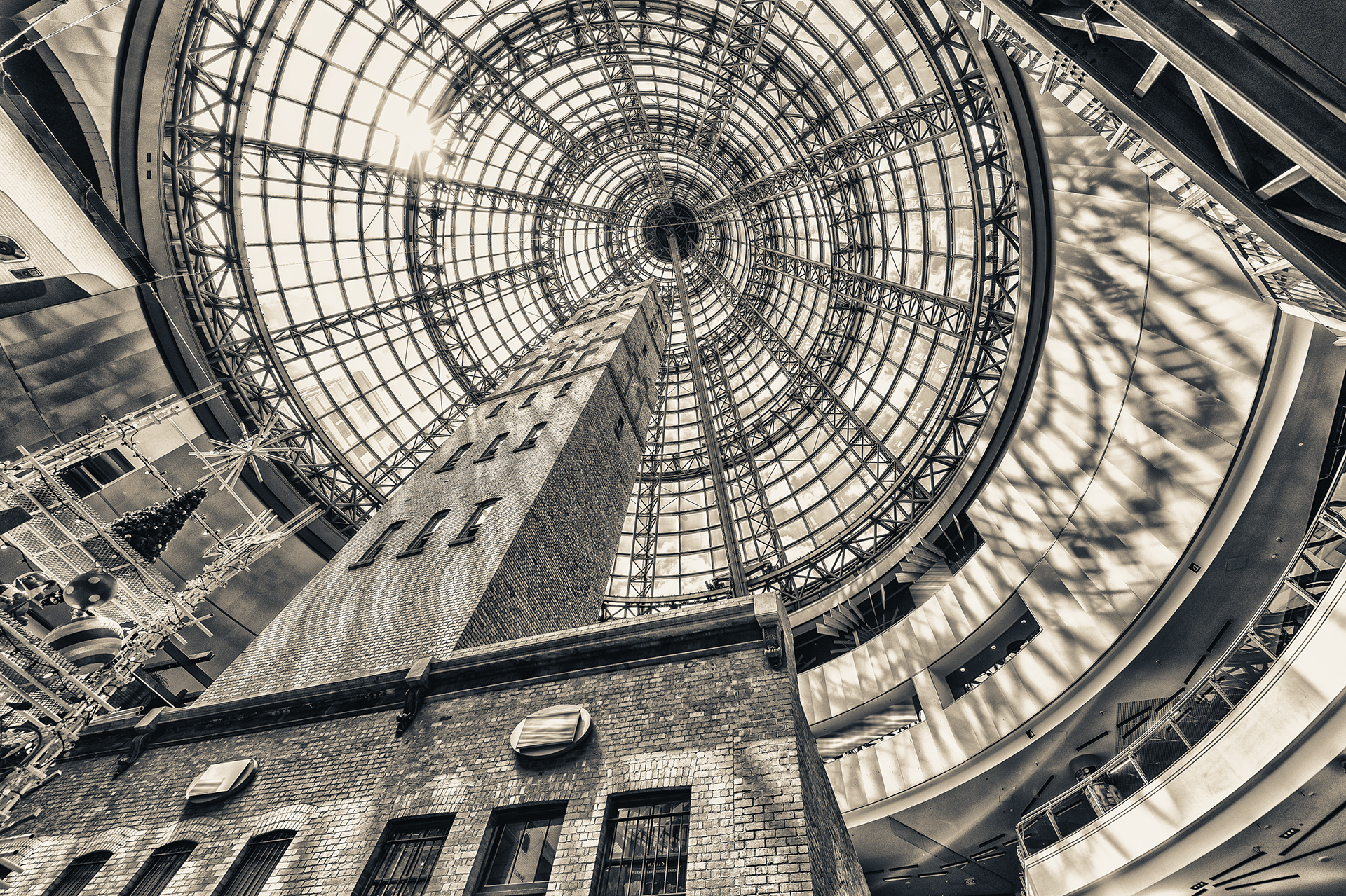 Black and white photograph looking upward inside Melbourne Central, showing the historic Shot Tower enclosed beneath the iconic glass dome roof, representing facility management and building operations in complex commercial environments.